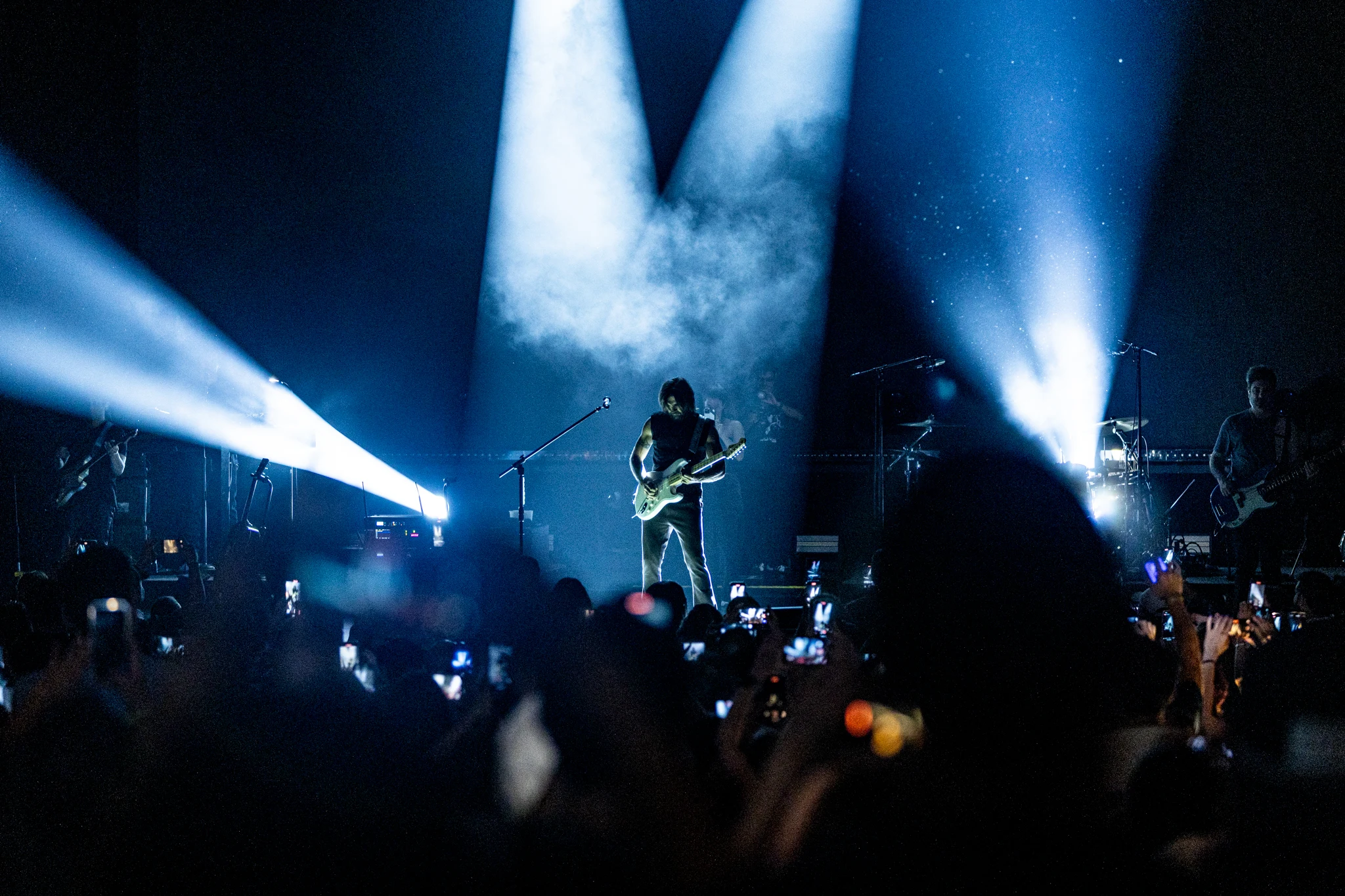 Guitarist performing solo under blue spotlights with stage smoke, crowd with phones raised at a LIVEST show.