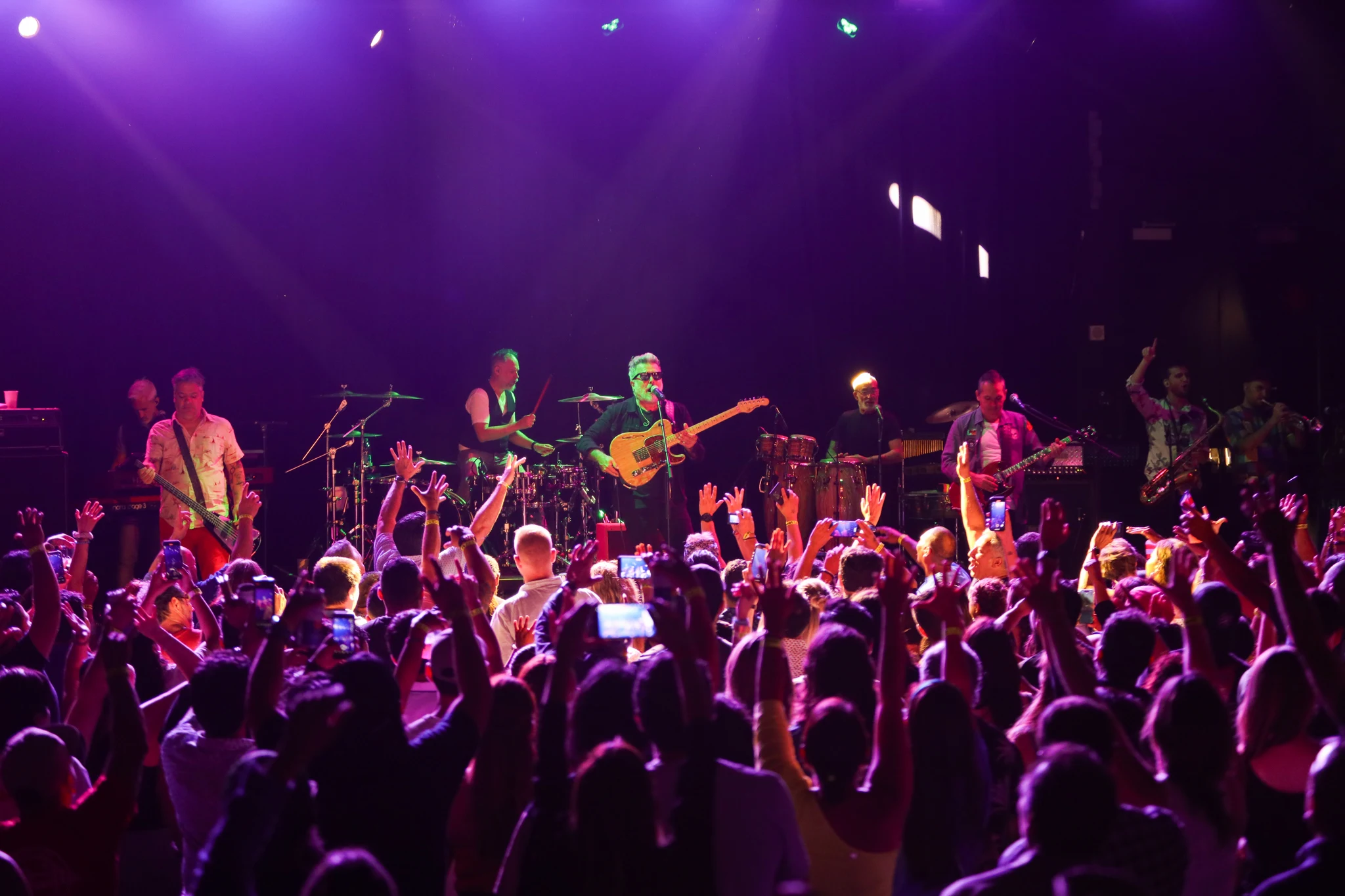 Band performing on stage with purple and green lighting, crowd with hands raised and phones out at a LIVEST show.
